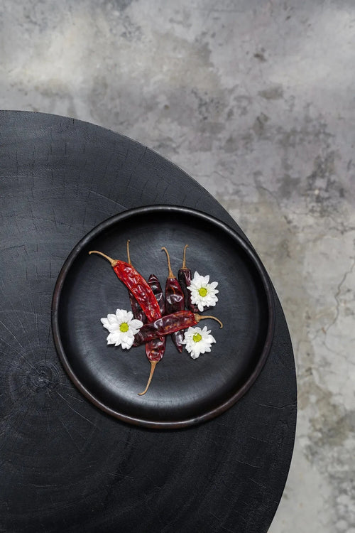 Assiette ronde en terre cuite brûlée noir profond Ø26 cm avec piments rouges séchés et fleurs blanches