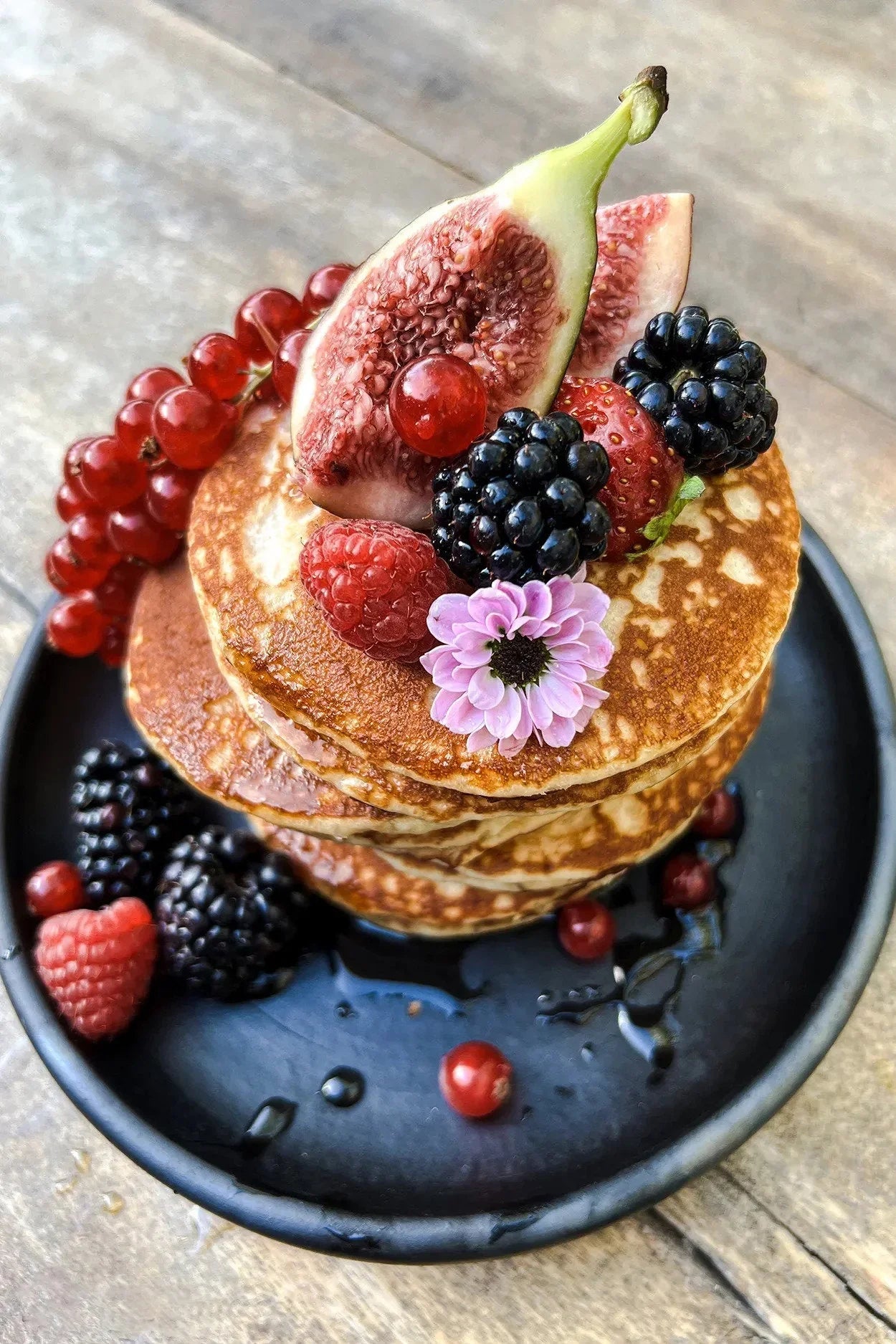 Assiette en terre cuite noire brûlée Ø20 cm avec pile de pancakes dorés, figues, baies et fleur violette