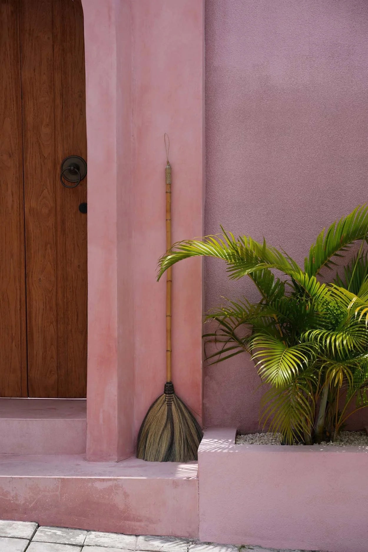 Balai décoratif en fibres naturelles beige et noir avec manche en bois courbé contre un mur rose