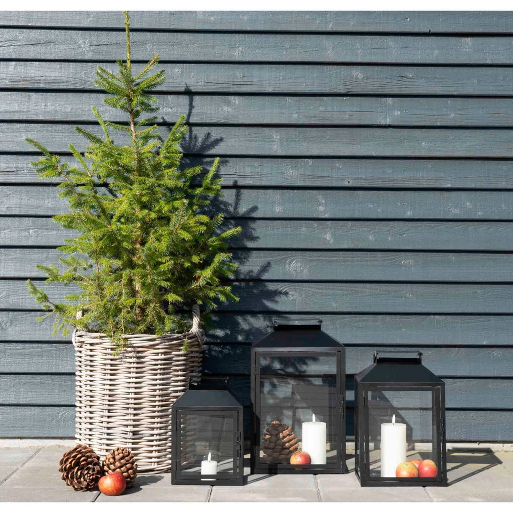 Lanterne en métal noir avec bougies blanches, entourées de pommes et pommes de pin