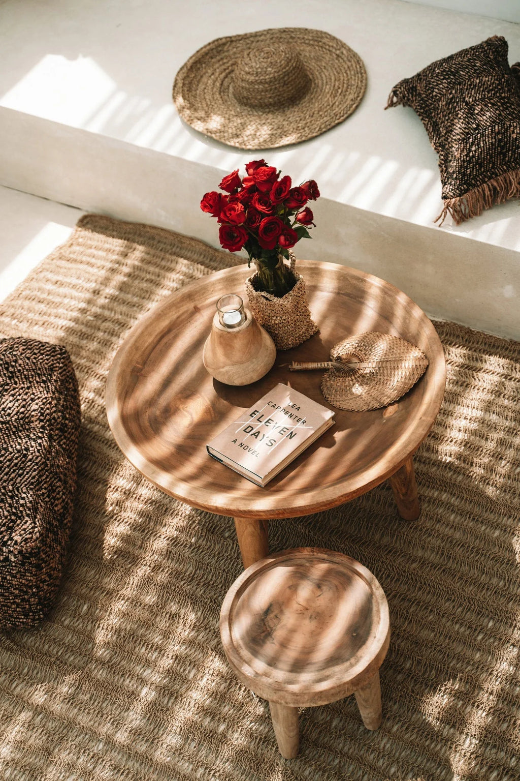 Table basse ronde en bois naturel sur 3 pieds, avec vase de roses rouges en panier tressé, bougeoir céramique et livre Ouvert Onze jours