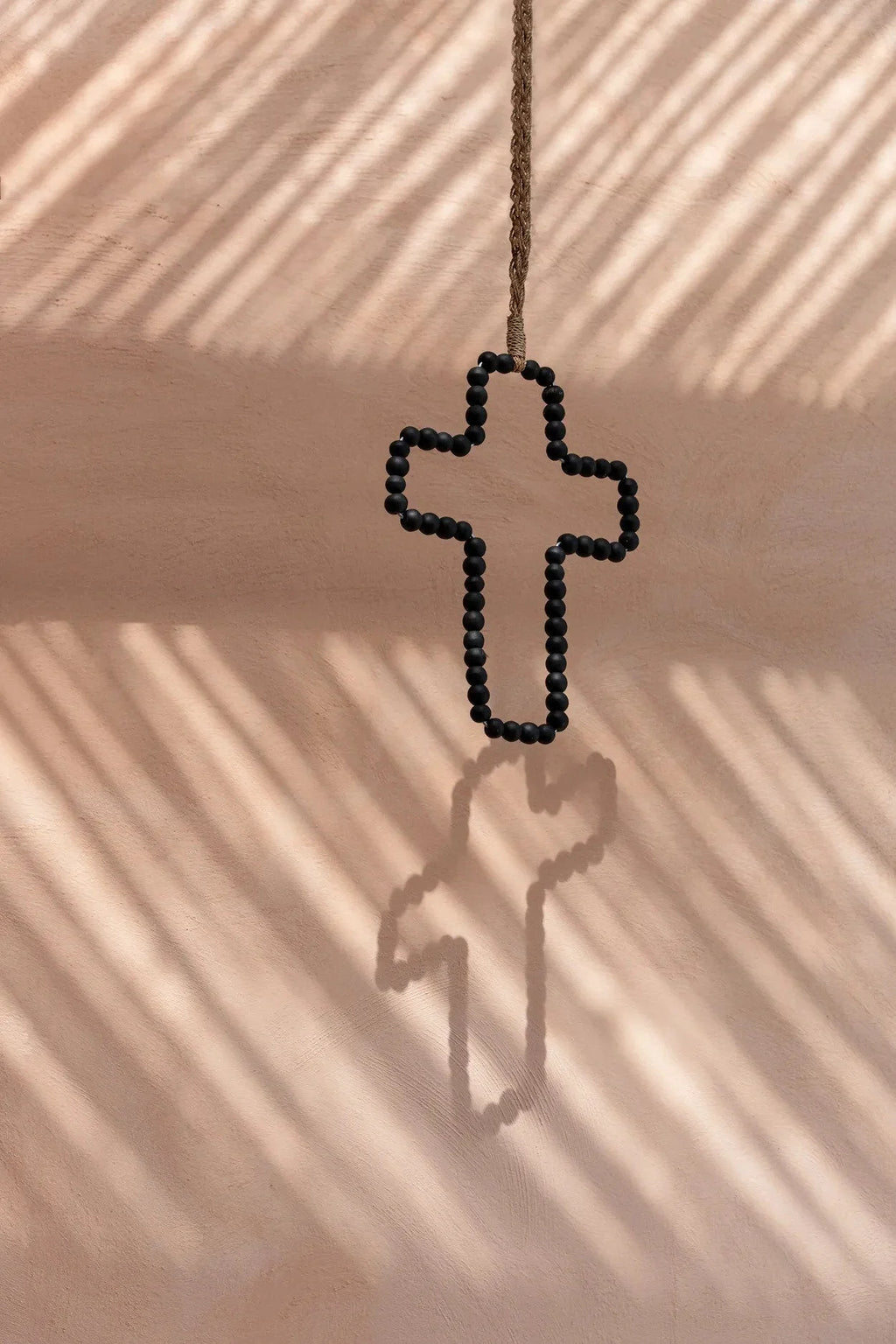 Croix murale décorative en bois noir perlé suspendue à une corde sur mur rose