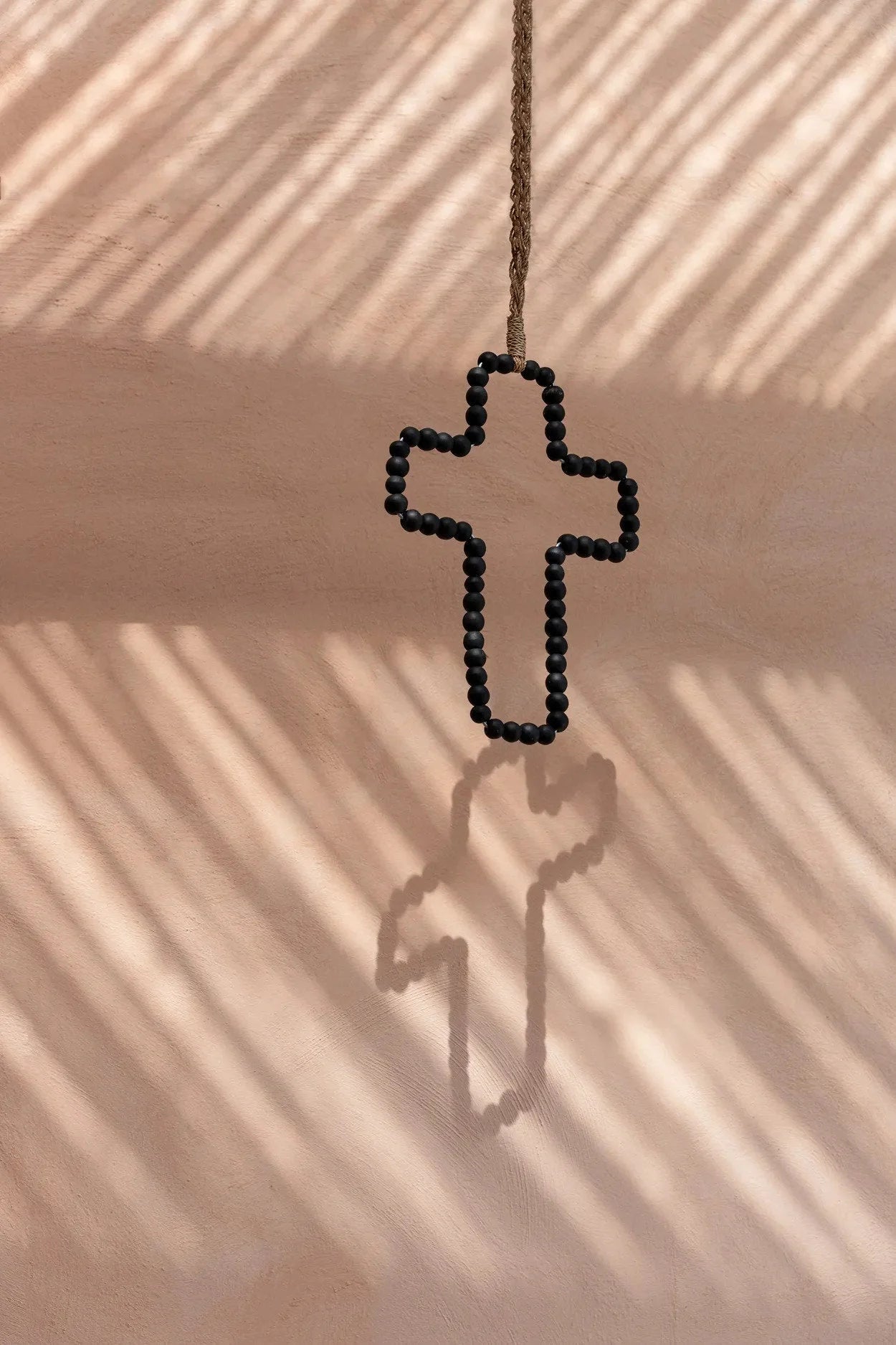 Croix murale décorative en bois noir perlé suspendue à une corde sur mur rose