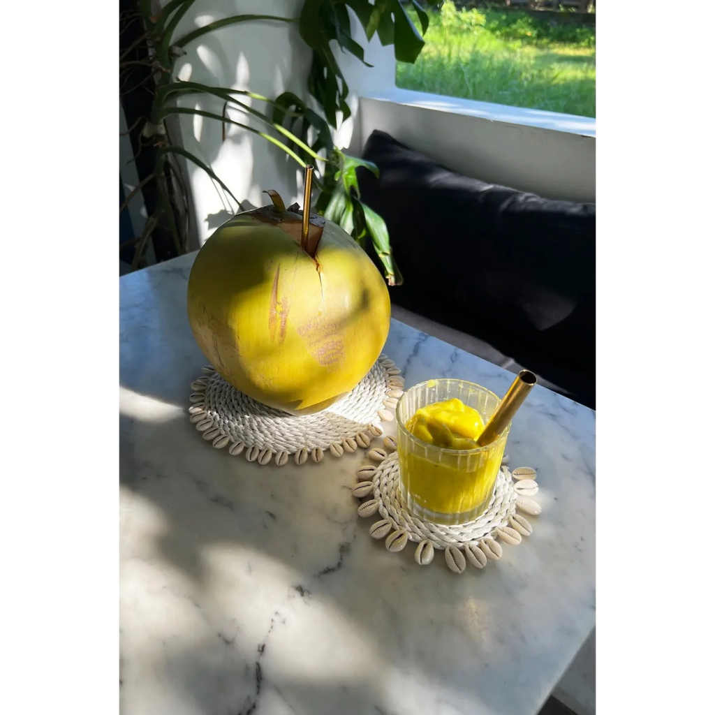 Coconut frais avec paille en métal et verre de boisson glacée jaune sur dessous de verre rond en herbe marine et coquillages