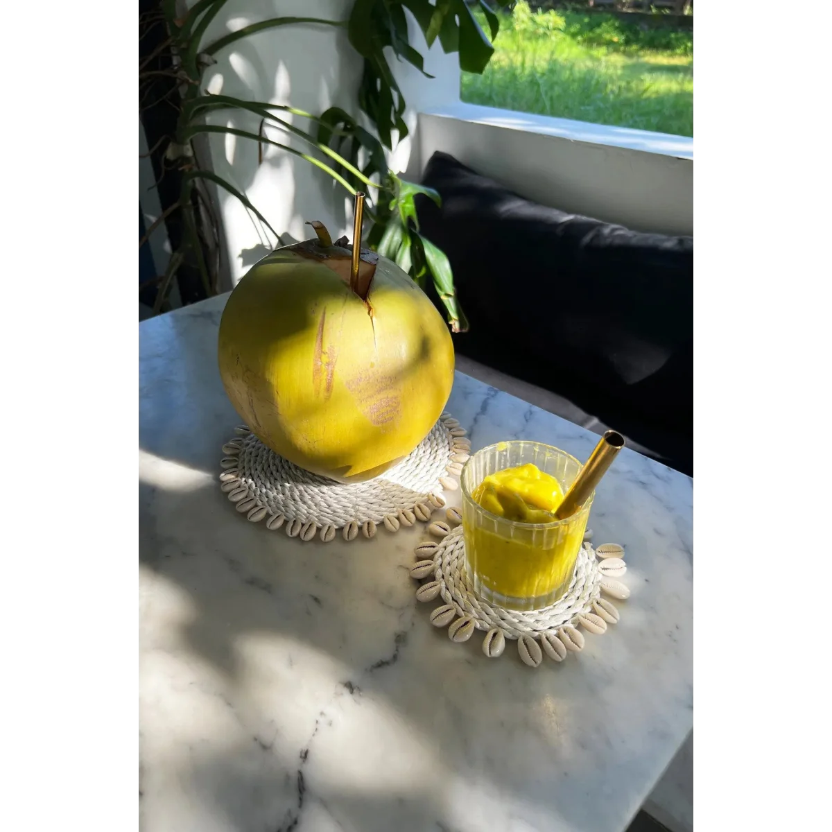 Coconut frais avec paille en métal et verre de boisson glacée jaune sur dessous de verre rond en herbe marine et coquillages