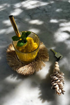 Verre de jus d’ananas jaune avec menthe et paille dorée sur sous-verre rond en raphia naturel à franges solaires