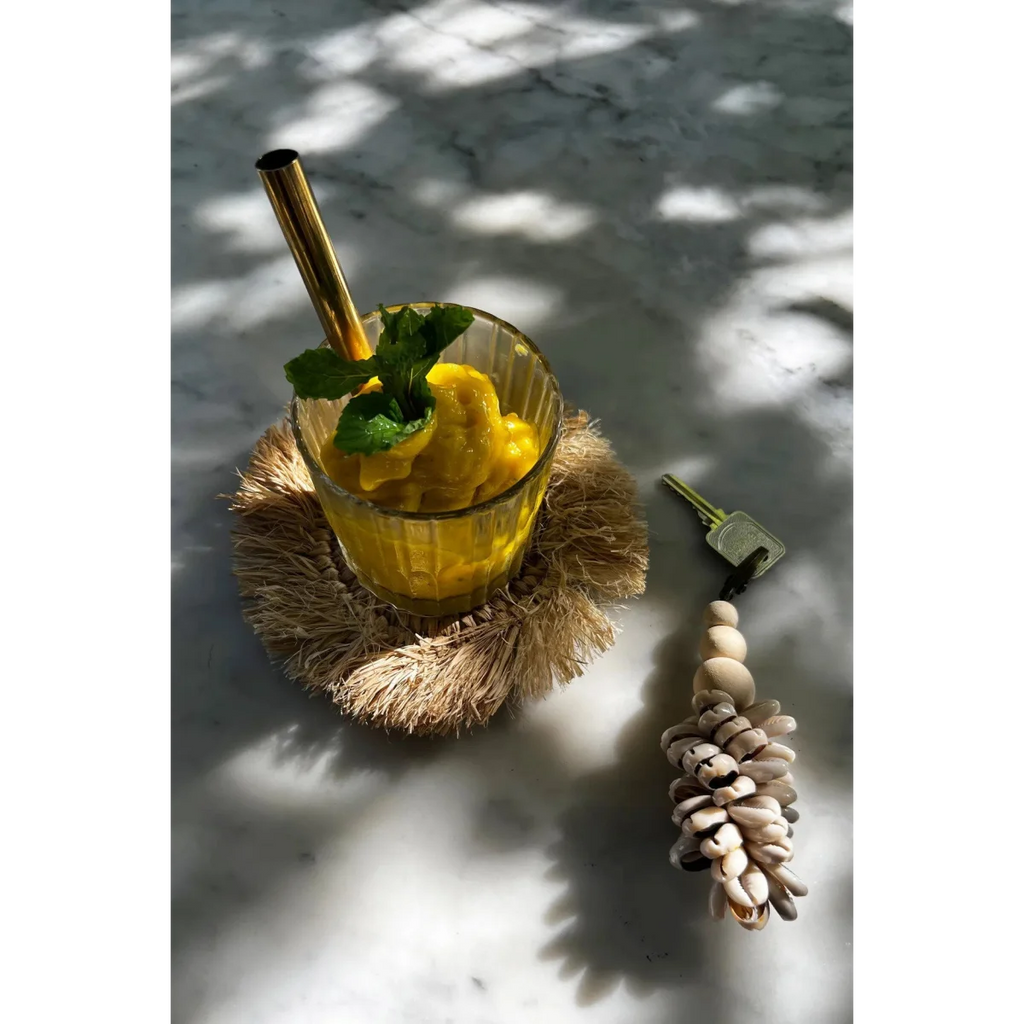 Verre de jus d’ananas jaune avec menthe et paille dorée sur sous-verre rond en raphia naturel à franges solaires