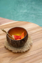 Verre en coque de noix de coco brun avec tranches d’orange et paille sur sous-verre rond en raphia naturel à franges solaires