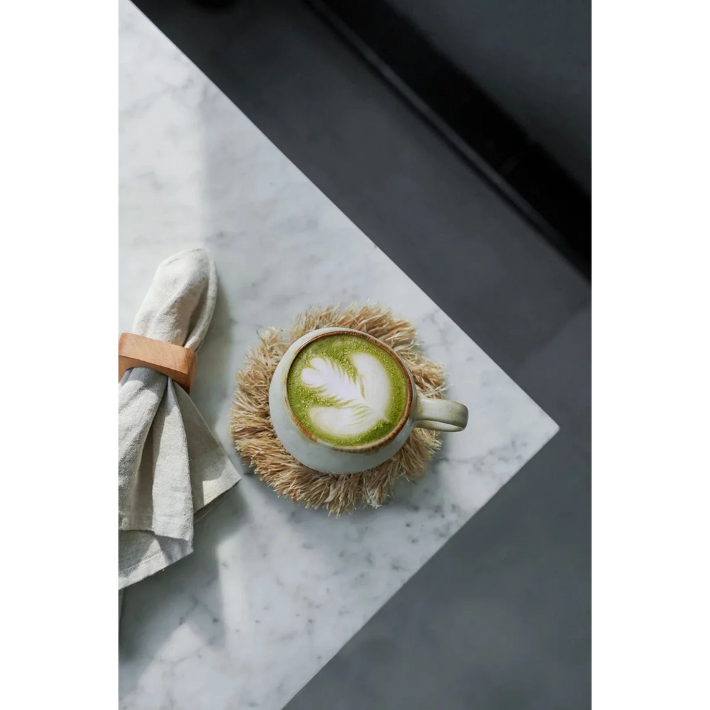 Tasse en céramique blanche pour matcha latte avec motif feuille de thé vert sur mousse, sur dessous de verre rond en raphia naturel à franges solaires
