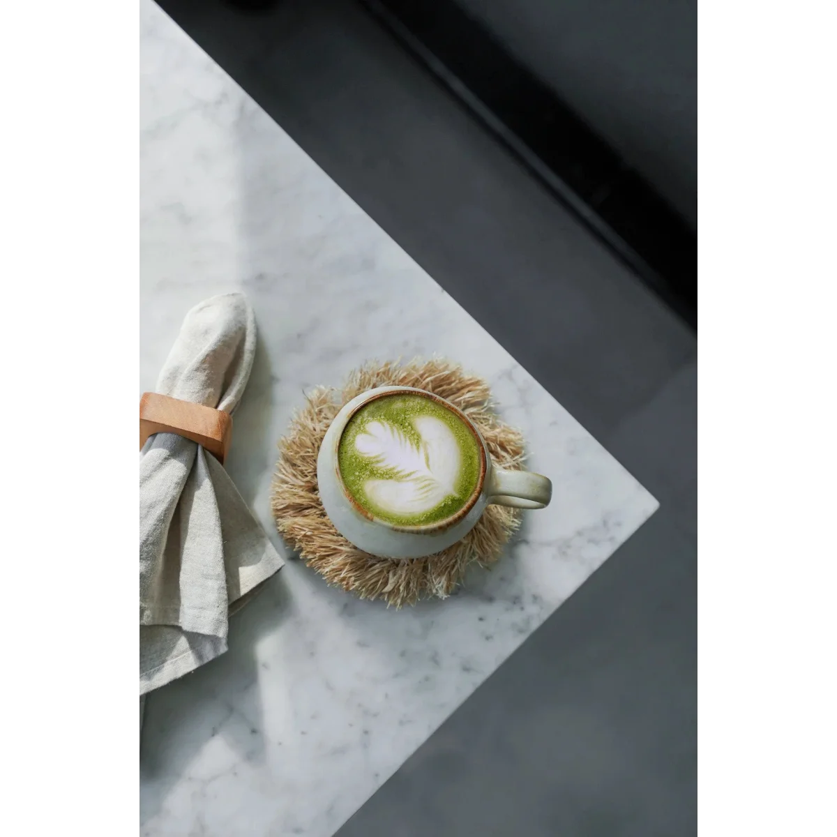 Tasse en céramique blanche pour matcha latte avec motif feuille de thé vert sur mousse, sur dessous de verre rond en raphia naturel à franges solaires