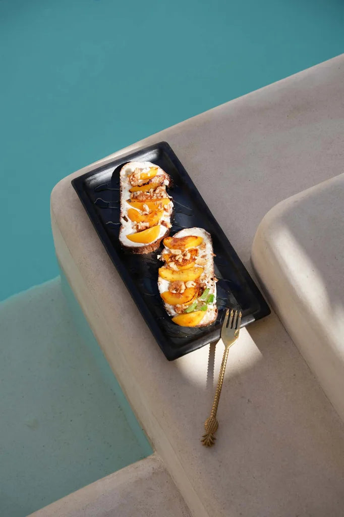 Toast de pêche et ricotta sur une assiette noire avec La Fourchette à Ananas Or