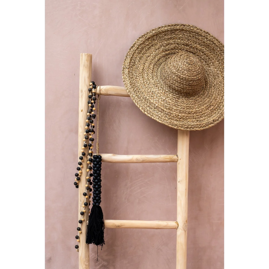 Chapeau paille sombrero avec perles noires et pompon sur échelle boisée contre mur rose