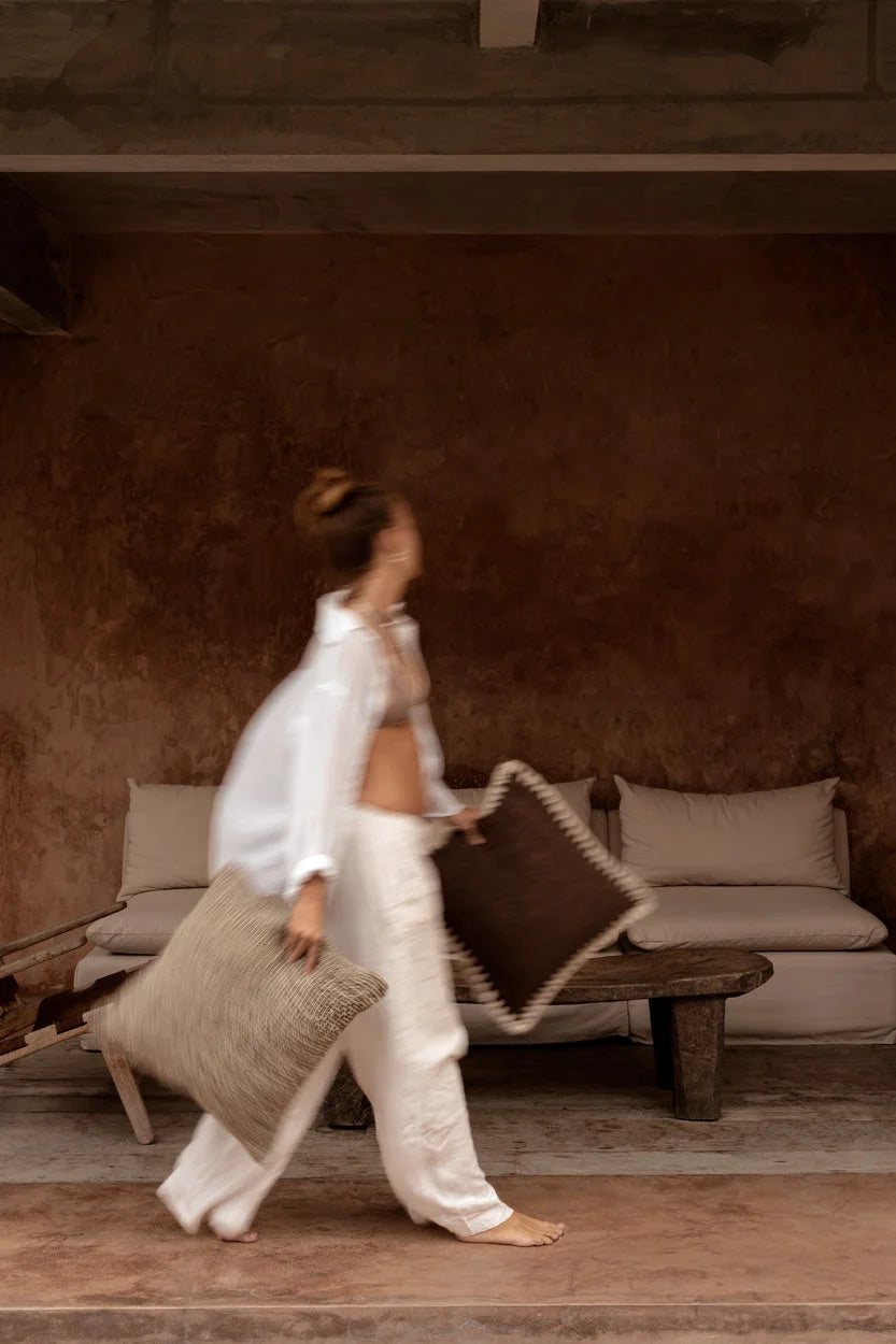 Femme en blanc portant deux coussins : beige texturé et marron à bord crocheté crème