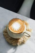 Tasse en céramique crème avec anse large et bord brun dégradé, latte art sur socle beige frangé