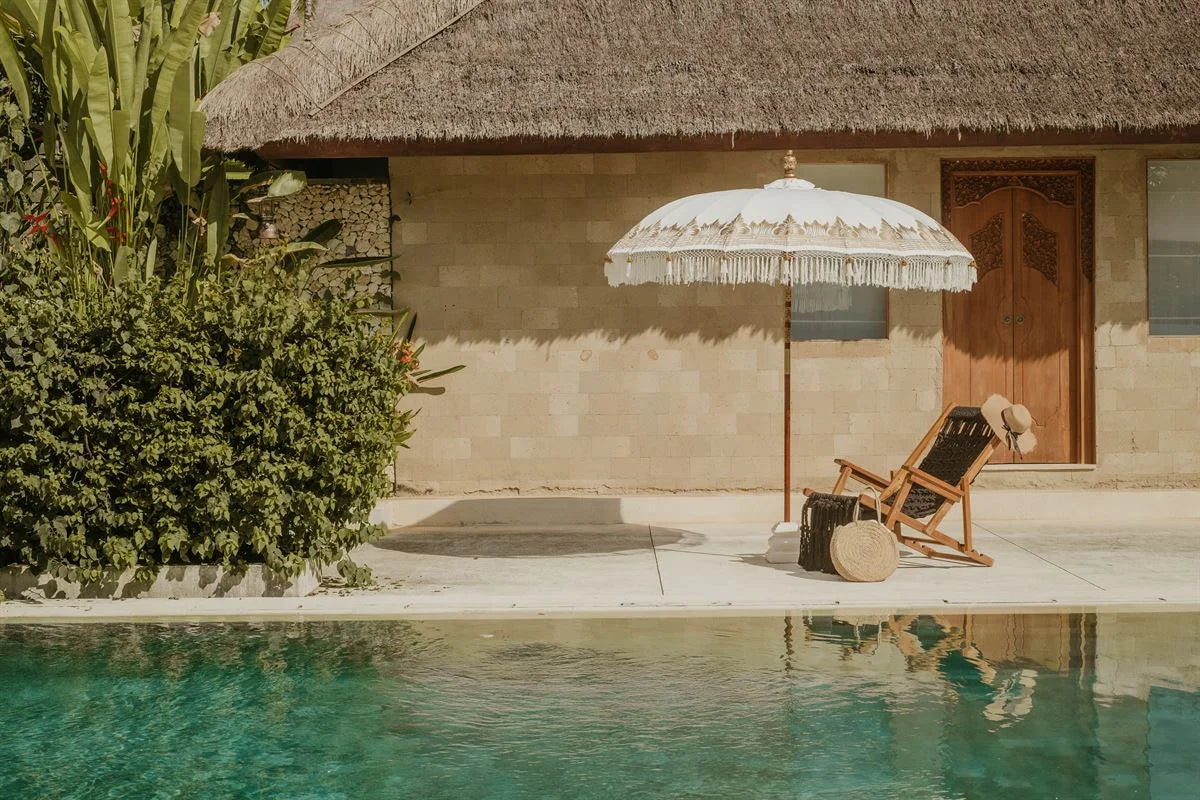 Coin lounge au bord de la piscine avec toiture en chaume et parasol crème à franges