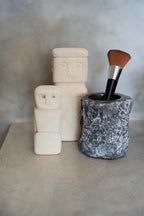 Deux figures stylisées crème avec un pot gris textured contenant un pinceau de maquillage