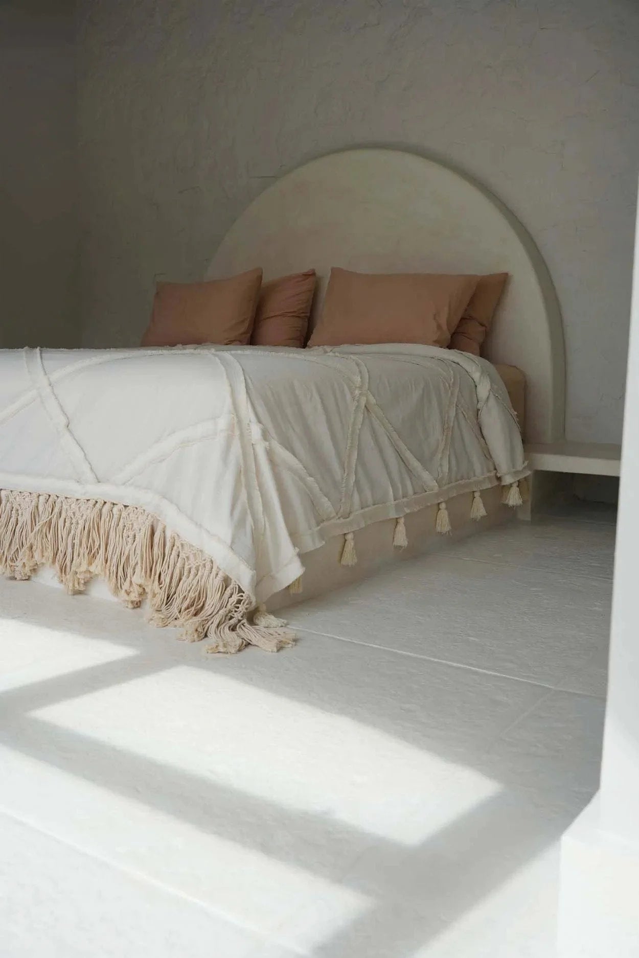 Lit blanc avec tête arrondie, coussins rose blush et plaid en coton écru à franges