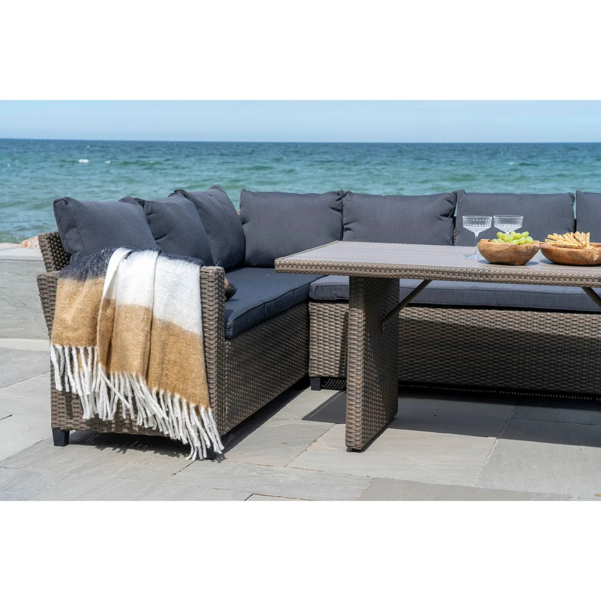 Canapé d’extérieur en rotin gris tissé avec coussins gris foncé et plaid frangé camel anthracite