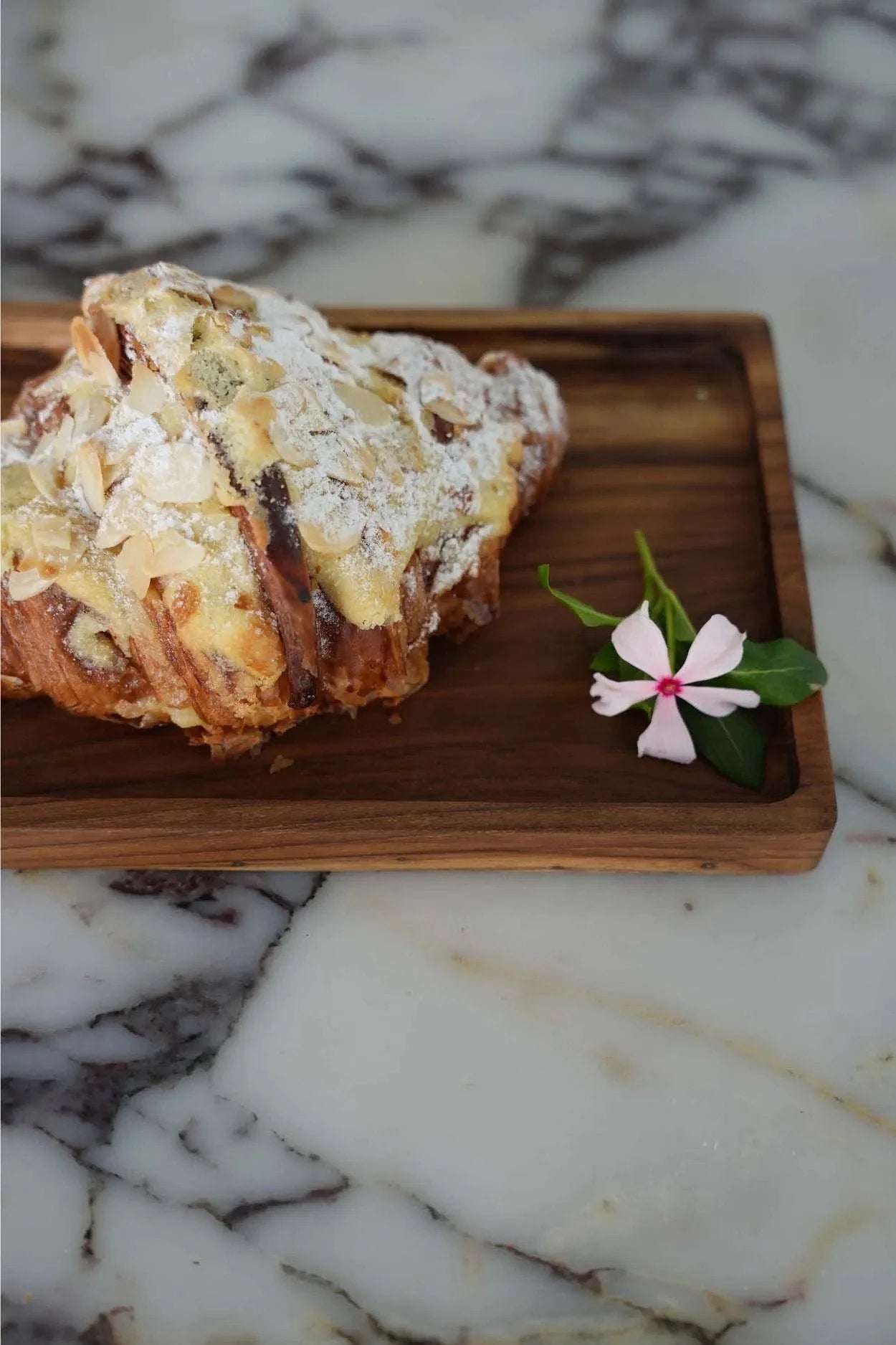 Croissant aux amandes saupoudré de sucre glace avec petite fleur rose sur Plateau en teck