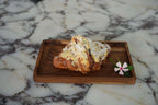 Croissant aux amandes saupoudré de sucre glace et décoré d’une petite fleur sur plateau en teck