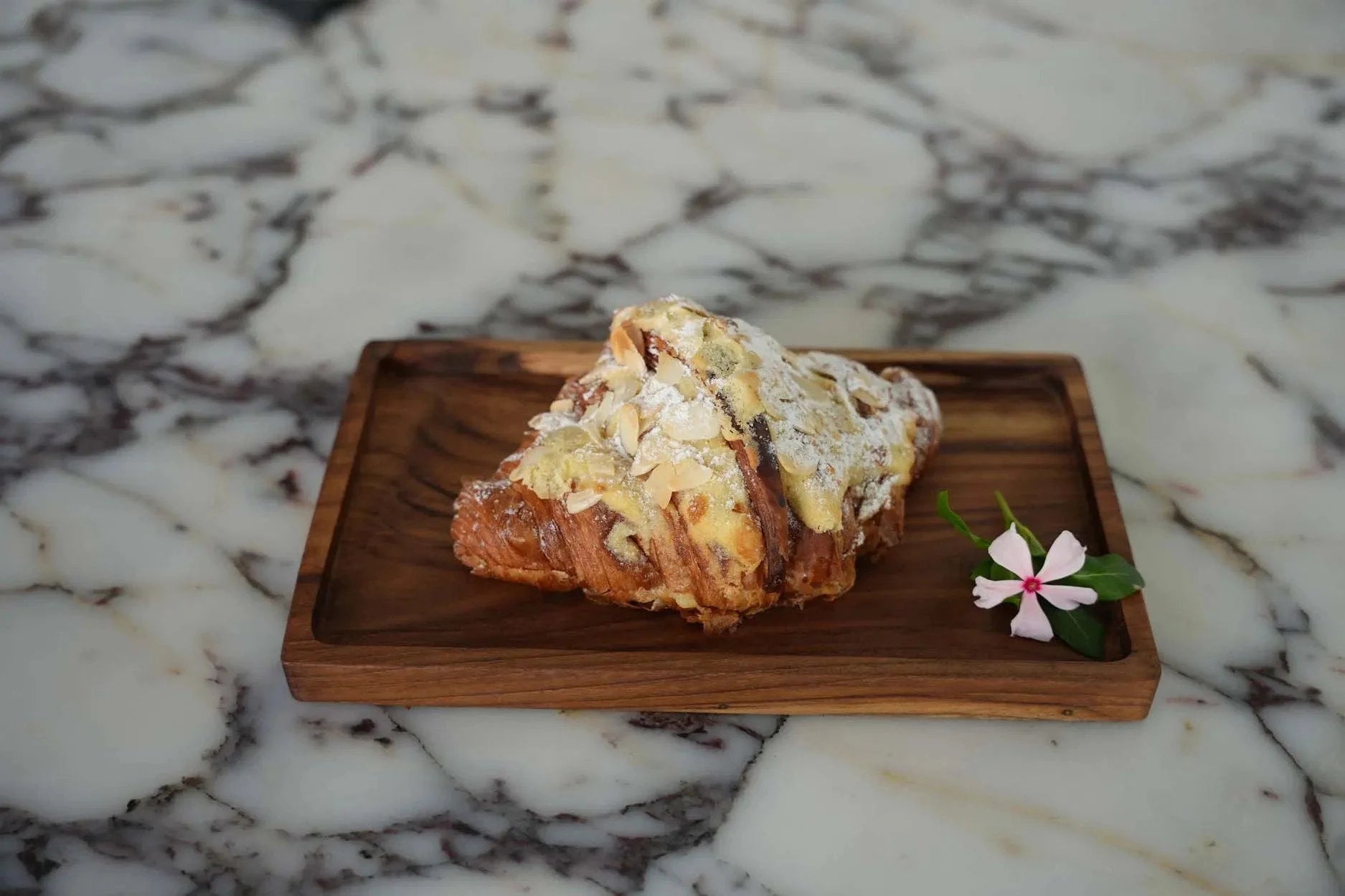 Croissant aux amandes saupoudré de sucre glace et décoré d’une petite fleur sur plateau en teck