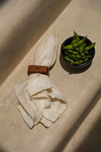 Edamame dans un bol foncé à côté d’une serviette en lin blanc avec un rond de serviette marron.