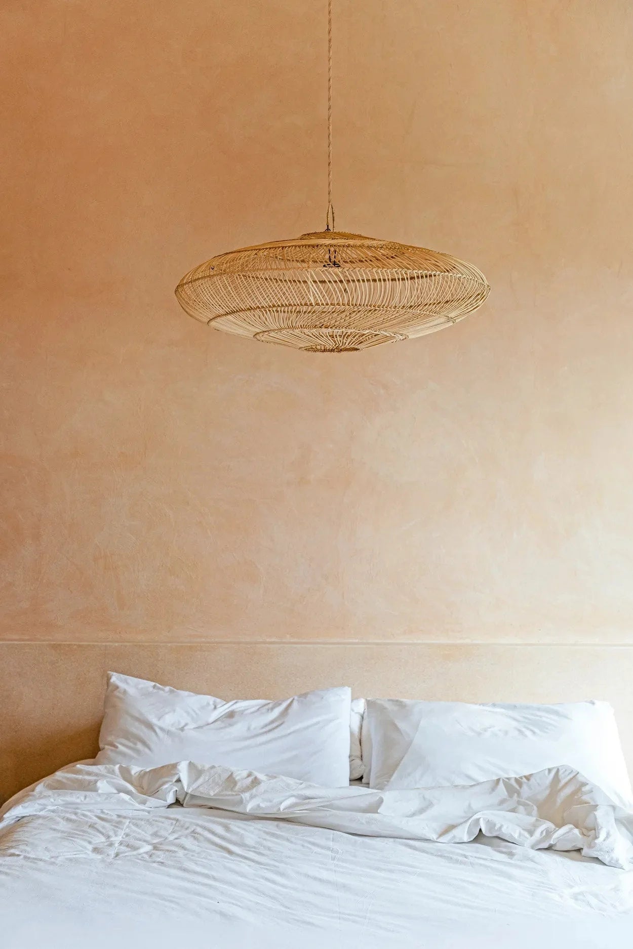 Suspension en rotin naturel forme soucoupe ajourée au-dessus d’un lit en lin blanc