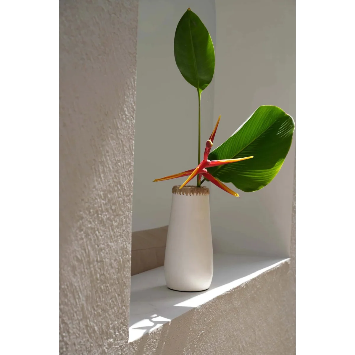 Vase en terracotta blanc naturel avec détail raphia, contenant deux feuilles vertes et une fleur rouge-orange